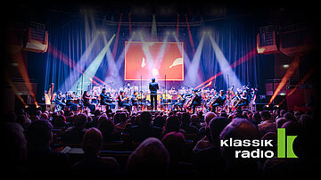 Menschen auf Stühlen blicken auf eine beleuchtete Bühne mit Orchester