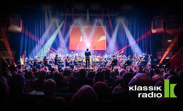 Menschen auf Stühlen blicken auf eine beleuchtete Bühne mit Orchester 