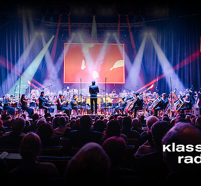 Menschen auf Stühlen blicken auf eine beleuchtete Bühne mit Orchester 