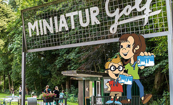 Miniatur Minigolfanlage mit Bäumen im Hintergrund. Im Vordergrund sind zwei Kinder mit einer swa City Karte. Weiterverlinkung auf swa City Minigolf in Wellenburg bei Augsburg Seite