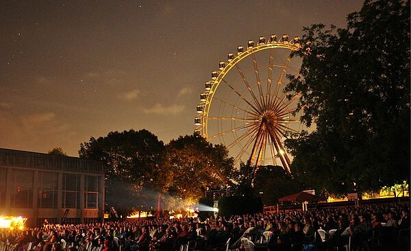 Viele Personen auf Stühlen bei der swa Kurzfilmnacht 2024. Im Hintergrund sind Bäume und ein Riesenrad