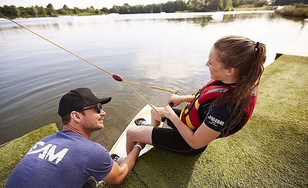 Frau steht auf einem Wakeboard im Wasser. Links neben ihr unterstützt sie ein Mann.