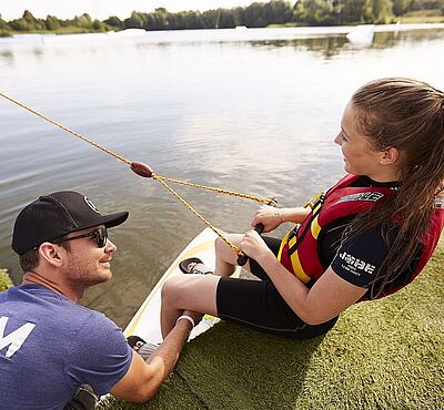 Frau steht auf einem Wakeboard im Wasser. Links neben ihr unterstützt sie ein Mann.
